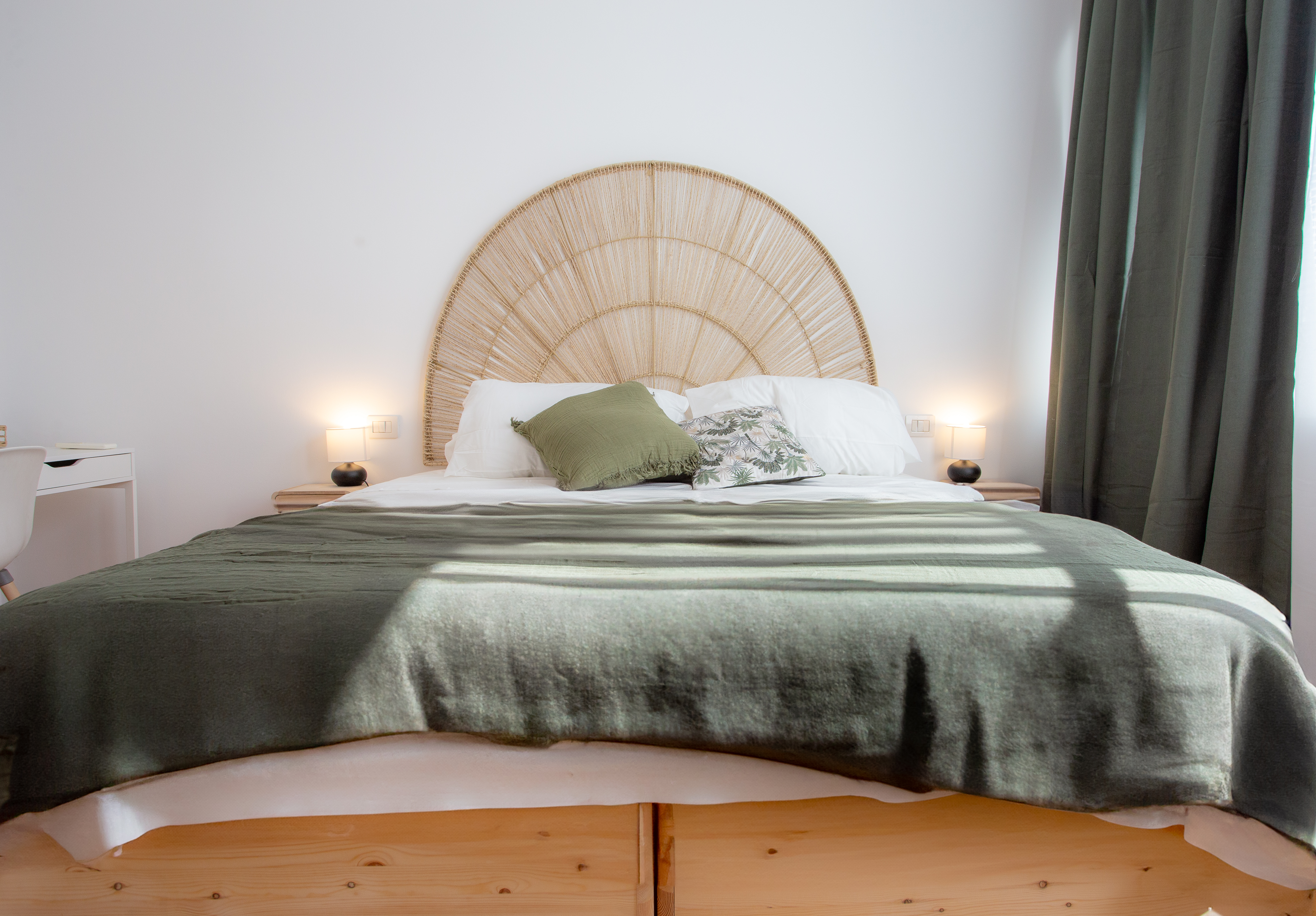 Cozy bedroom featuring a large rattan headboard, green bedding, and decorative pillows, with a minimalist white wall and natural light.